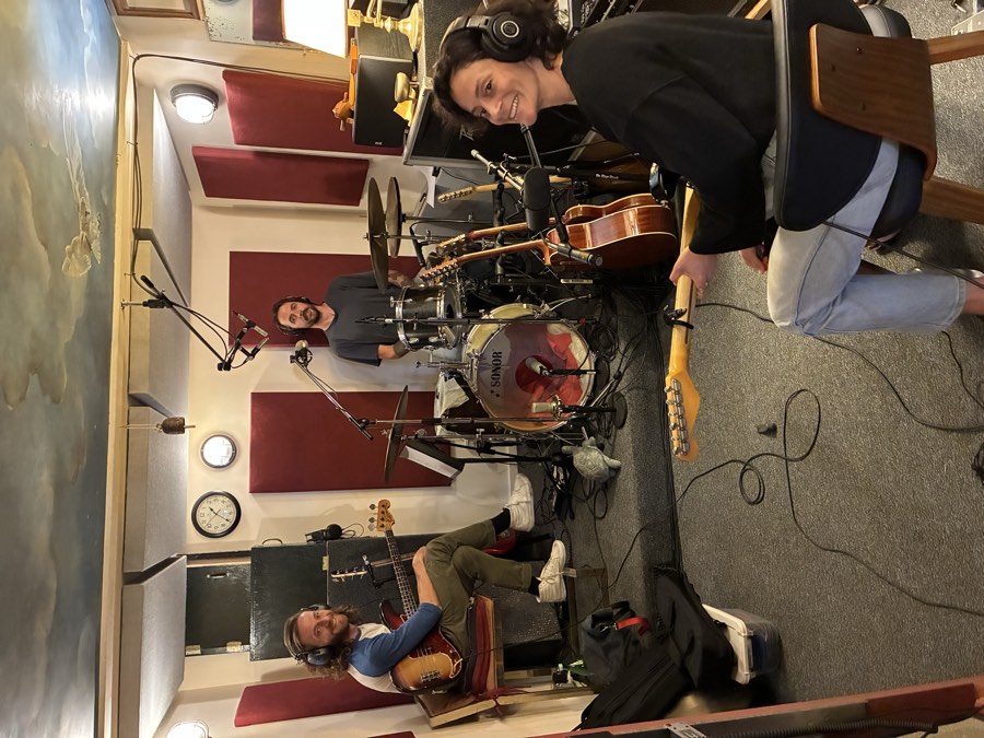 Three musicians tracking together in the live room, playing bass, drums and guitar with headphones and microphones set up