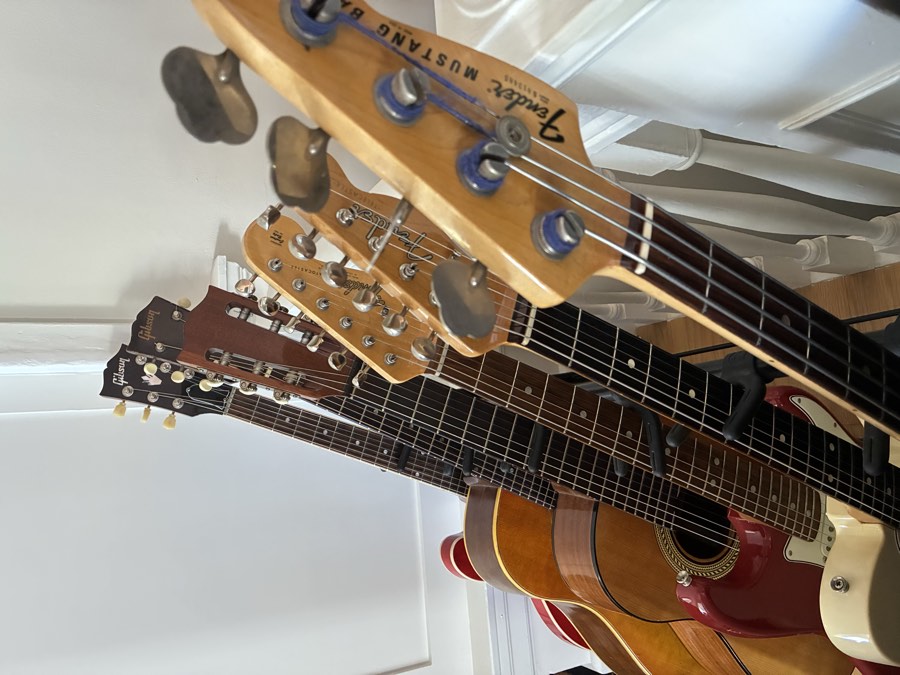 Vintage guitars and basses, including Gibson acoustics and a Fender Mustang Bass, lined up on a guitar stand